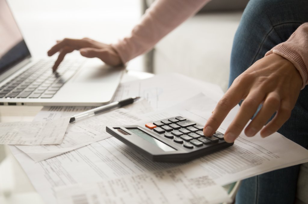 Close up cropped image young woman calculating monthly expenses, managing budget, entering data in computer application, sitting at table full of papers, loan documents, invoices, utility bills.